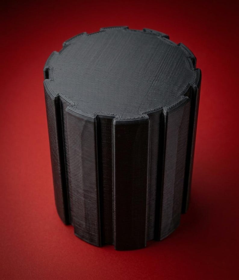 K&ouml;nnte beinhalten: Ein schwarzes zylindrisches Objekt mit einer zahnradartigen, gekerbten Oberkante und vertikalen Rippen an den Seiten. Die Oberfl&auml;che weist durchgehend feine, horizontale 3D-Druckschichten auf, die eine texturierte, matte Oberfl&auml;che erzeugen.