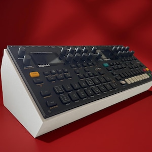 May include: A black Elektron Digitakt drum computer and sampler mounted on a white angled stand. The device features a dark interface with numerous knobs, a small screen, and a grid of backlit buttons labelled with numbers and various control functions.