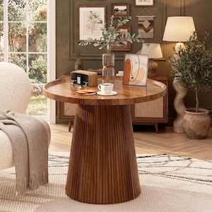 May include: A round wooden side table with a fluted, pedestal-style base. The tabletop holds a clear glass vase with green foliage, a small white ceramic cup on a saucer, a wooden radio, and a framed abstract line art print.