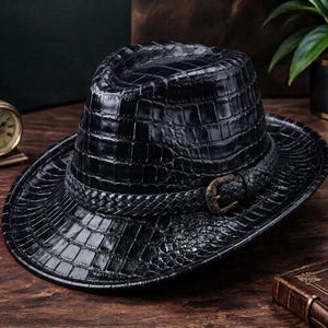 Op de afbeelding: Een zwarte fedora hoed met een gestructureerd krokodillenleerpatroon over de kroon en de rand. De hoed is geaccentueerd met een gevlochten zwarte band die rond de basis loopt, vastgezet met een metalen gesp met een antieke bronzen afwerking.