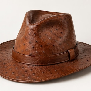 Peut inclure: Un chapeau fedora marron doté d'une surface texturée avec de petites indentations sombres régulièrement espacées. Le chapeau comprend une bande marron assortie entourant la base de la calotte, avec un sommet pincé et un bord large.