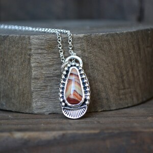 May include: A silver necklace with a teardrop-shaped pendant featuring a red, white, and brown agate stone. The pendant is surrounded by a beaded border and has a decorative design at the bottom.