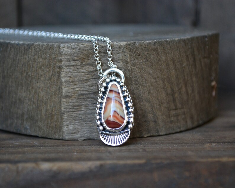 May include: A silver necklace with a teardrop-shaped pendant featuring a red, white, and brown agate stone. The pendant is surrounded by a beaded border and has a decorative design at the bottom.