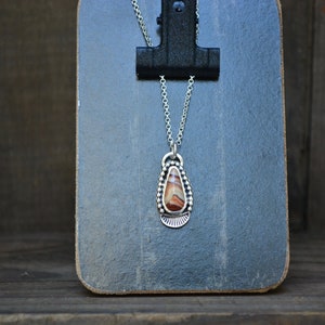 May include: A silver necklace with a teardrop-shaped pendant featuring a brown and white agate stone. The pendant is surrounded by a beaded border.