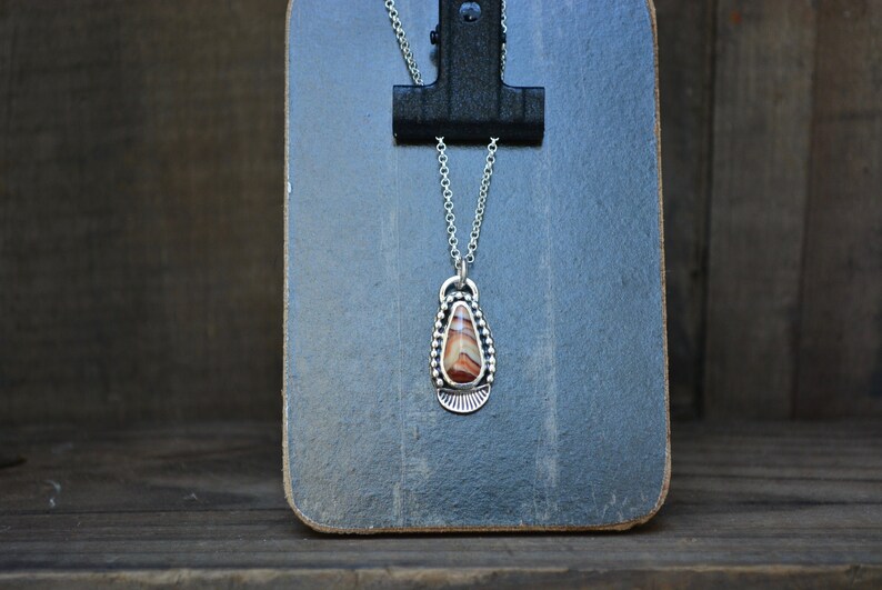 May include: A silver necklace with a teardrop-shaped pendant featuring a brown and white agate stone. The pendant is surrounded by a beaded border.