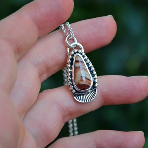 May include: A silver necklace with a teardrop-shaped pendant featuring a brown and white agate stone. The pendant is surrounded by a beaded border and a decorative silver design.