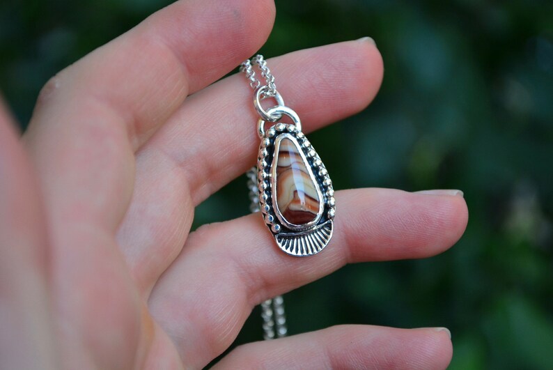 May include: A silver necklace with a teardrop-shaped pendant featuring a brown and white agate stone. The pendant is surrounded by a beaded border and a decorative silver design.