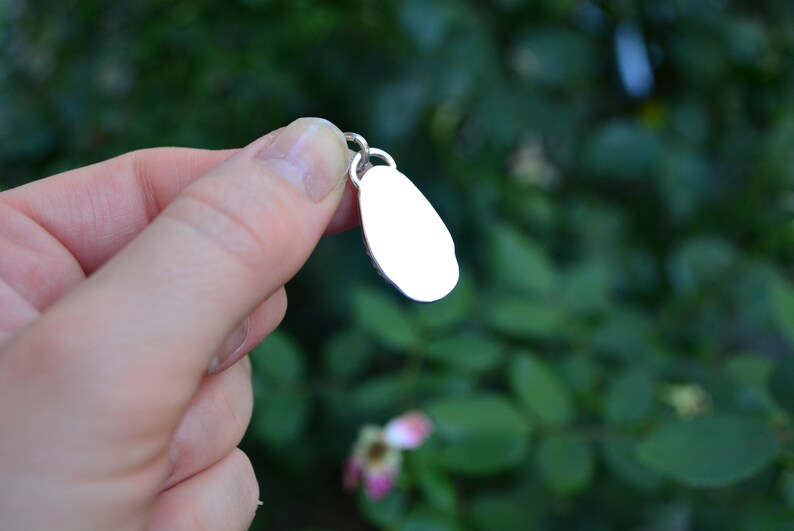 May include: A silver charm in the shape of an oval, with a smooth, polished surface. The charm is hanging from a silver loop.