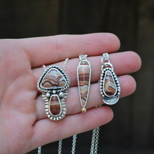 May include: Three silver necklaces with brown and white agate stone pendants. The pendants are different shapes, including a teardrop, a triangle, and a mushroom shape.