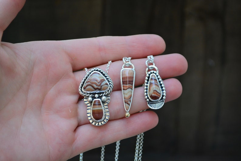 May include: Three silver necklaces with brown and white agate stone pendants. The pendants are different shapes, including a teardrop, a triangle, and a mushroom shape.