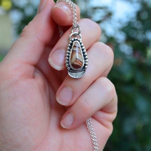 May include: A silver pendant necklace with a teardrop-shaped stone featuring brown and white stripes. The stone is set in a silver bezel with a beaded border.