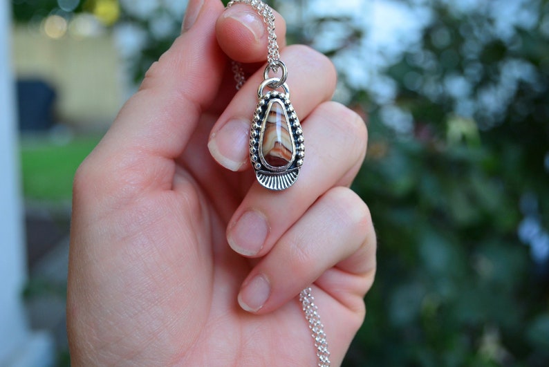 May include: A silver pendant necklace with a teardrop-shaped stone featuring brown and white stripes. The stone is set in a silver bezel with a beaded border.