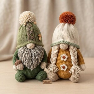 May include: Two crocheted gnome dolls. The left gnome wears a green hat with a cream pom-pom, a cable knit detail, and a small mushroom charm, featuring a gray beard and holding an acorn. The right gnome has a cream hat with a rust pom-pom, two long blonde braids, and a mustard yellow body with white flower appliqués.