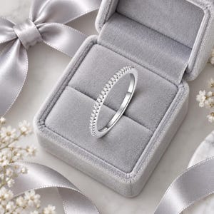 May include: A thin silver-toned band ring featuring a row of small, clear, round-cut stones set along the top half. The ring is displayed inside a square, light gray velvet-lined jewelry box with a matching gray satin ribbon tied around it.