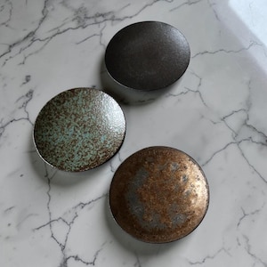 May include: Three circular plates with distinct finishes. One plate has a dark charcoal gray surface, another features a mottled teal and brown speckled glaze, and the third displays a textured metallic bronze and dark gray patina finish.