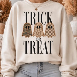 Puede incluir: Una sudadera de cuello redondo color crema con el texto negro TRICK OR TREAT y tres ilustraciones de fantasmas. Los fantasmas tienen diferentes patrones: uno de camuflaje, uno de estampado de leopardo y otro de cuadros marrones y crema.