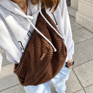 May include: A brown corduroy crossbody bag with a textured ribbed surface and a small rectangular brown leather-like tag featuring white text. The bag is worn over a light gray hooded sweatshirt with black lettering and light blue denim jeans.