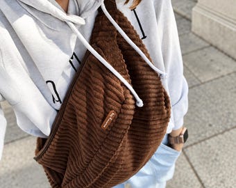 Peut inclure: Un sac bandoulière en velours côtelé marron avec une surface texturée à côtes et une petite étiquette rectangulaire en similicuir marron portant du texte blanc. Le sac est porté sur un sweat à capuche gris clair et un jean bleu clair.