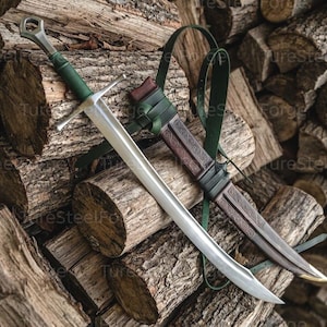 Puede incluir: Una espada de acero con empuñadura forrada en cuero verde y una guarda, acompañada de una vaina de cuero marrón con patrones de volutas en relieve y una correa de cuero verde, apoyada sobre una pila de troncos de madera.