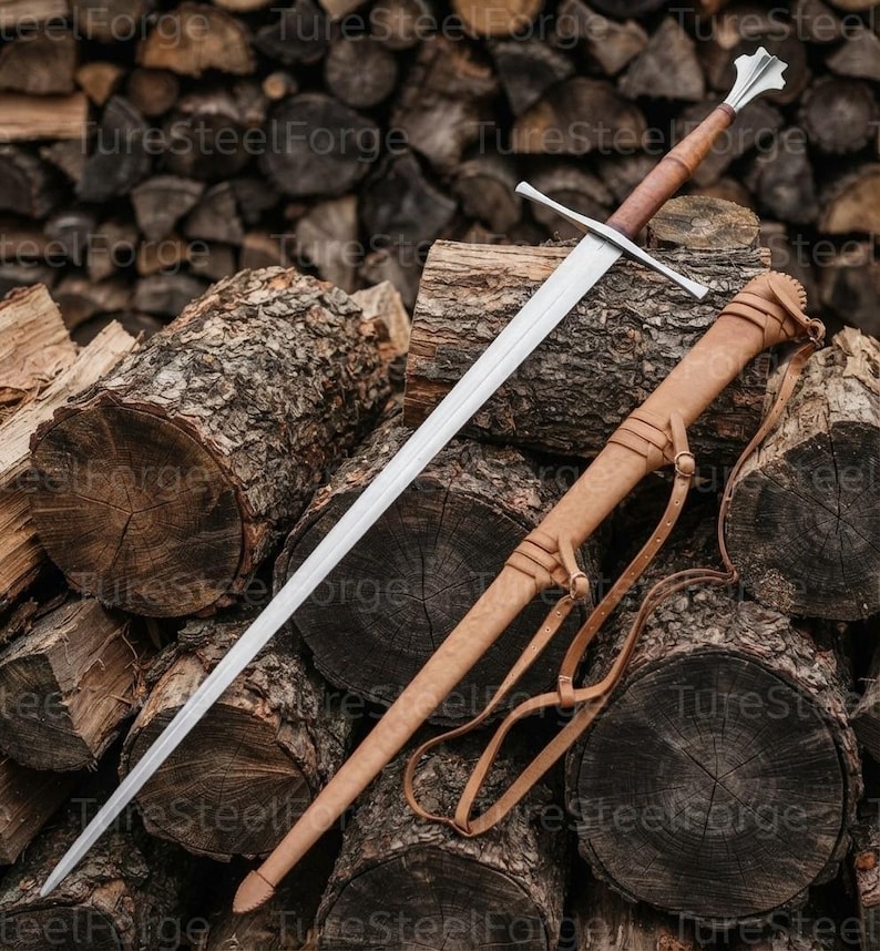 Puede incluir: Una espada larga de acero con hoja de doble filo, guarda recta y empu&ntilde;adura forrada en cuero marr&oacute;n que termina en un pomo met&aacute;lico. Junto a la espada hay una vaina de cuero color canela a juego con correas y hebillas integradas.