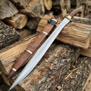 Puede incluir: Una espada de acero de doble filo con punta afilada y canal central, que presenta un mango de madera marrón oscuro con una guarda de latón. A su lado se encuentra una vaina de madera a juego con bandas de latón y una correa de cuero negro.