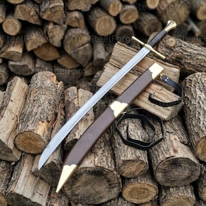 Può includere: Una spada in acciaio con lama dritta, guardia a croce e impugnatura scura, accompagnata da un fodero in pelle marrone con finiture metalliche color ottone sulla punta e sull'imboccatura, con cinghie in pelle nera per il trasporto.