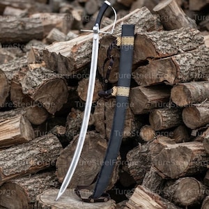 Puede incluir: Un sable de acero curvado con mango negro y guarda plateada, acompañado de una funda de cuero negro a juego con bandas decorativas de color dorado y una fina correa de cuero con pequeños remaches metálicos para llevar o asegurar la hoja.