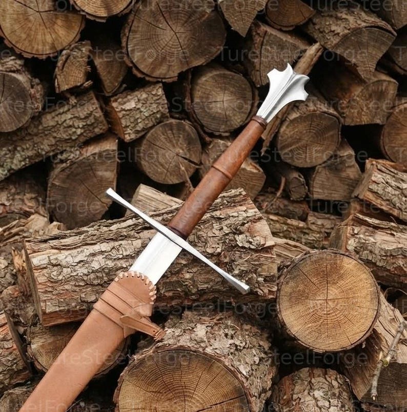 Puede incluir: Una espada larga con hoja de metal plateado, guarda recta y empu&ntilde;adura envuelta en cuero marr&oacute;n. El pomo presenta un dise&ntilde;o estriado de varios l&oacute;bulos. La parte inferior de la hoja est&aacute; encajada en una vaina de cuero color canela con costuras.