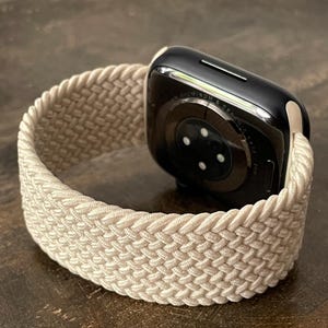 May include: A smartwatch with a black casing and a textured, braided sand-colored elastic band. The back of the watch features a circular sensor array and the text Sand is printed in the bottom left corner of the image.