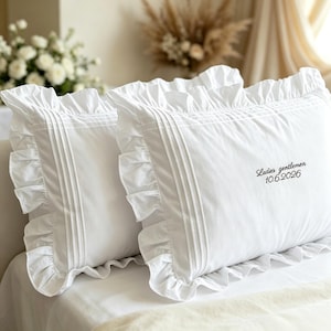 Puede incluir: Dos fundas de almohada rectangulares blancas con bordes de volantes y pliegues verticales. La funda derecha está bordada con el texto negro "Ladies gentlemen 10.6.2026" en una fuente cursiva, situada en el lado derecho.
