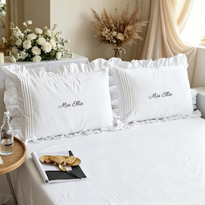 Puede incluir: Dos fundas de almohada rectangulares blancas con bordes con volantes y detalles de alforzas verticales. Cada funda está bordada con el nombre Miss Ellie en una fuente cursiva negra, centrada en la superficie de tela blanca lisa.