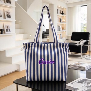 Può includere: Una borsa tote con un motivo a righe verticali blu navy e bianco, dotata di due lunghi manici a spalla e una chiusura a coulisse sulla parte superiore. Il nome Gavin è ricamato in corsivo viola al centro della parte anteriore.