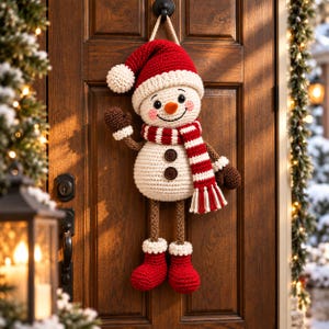 Puede incluir: Un adorno de pared de muñeco de nieve tejido a ganchillo con un gorro de Papá Noel rojo y blanco, una bufanda a rayas a juego, dos botones marrones en el torso, brazos con manoplas, botas rojas, una nariz de zanahoria y mejillas rosadas.