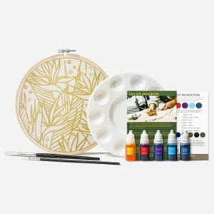 May include: A batik craft kit featuring a circular wooden embroidery hoop with a gold botanical print on fabric, a white plastic paint palette, two black paintbrushes, five small dye bottles labelled red, violet, cyan, navy blue, and yellow.