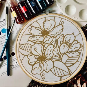 Könnte beinhalten: Ein runder Stickrahmen aus Holz mit weißem Stoff, der ein florales Linienmuster in Gold zeigt. Das Design besteht aus drei Blüten und Blättern. Zwei Pinsel, mehrere kleine Tintenfläschchen und eine weiße Farbpalette liegen daneben.