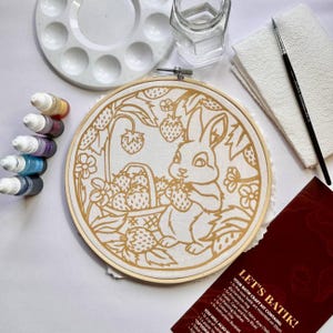 Könnte beinhalten: Ein Batik-Bastelset mit einem runden Bambusrahmen, der ein vorgedrucktes Hasen- und Erdbeermotiv auf Stoff enthält, sowie einer weißen Kunststoff-Farbpalette, einem Glaswasserbehälter, einem dünnen Pinsel und einer dunkelroten Anleitung.