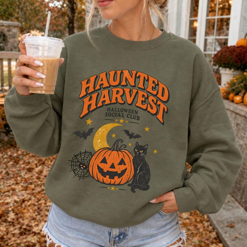 May include: An olive green crewneck sweatshirt featuring a graphic print with the text "HAUNTED HARVEST HALLOWEEN SOCIAL CLUB" in orange. The design includes a yellow crescent moon, a jack-o'-lantern, a spider web, bats, and a black cat.