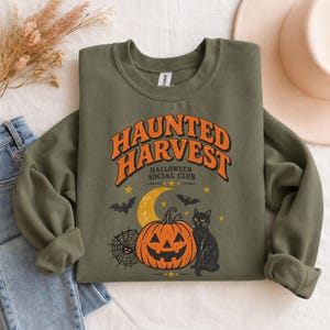 May include: An olive green crewneck sweatshirt featuring a graphic print with the text "HAUNTED HARVEST HALLOWEEN SOCIAL CLUB" in orange and black. The design includes a jack-o'-lantern, a black cat, a crescent moon, bats, and a spider web.