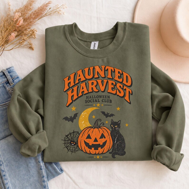 May include: An olive green crewneck sweatshirt featuring a graphic print with the text "HAUNTED HARVEST HALLOWEEN SOCIAL CLUB" in orange and black. The design includes a jack-o'-lantern, a black cat, a crescent moon, bats, and a spider web.