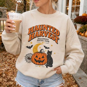 May include: A beige crewneck sweatshirt featuring a graphic print with the text "HAUNTED HARVEST HALLOWEEN SOCIAL CLUB" in orange and black. The design includes a jack-o'-lantern, a black cat, a crescent moon, bats, and a spider web.