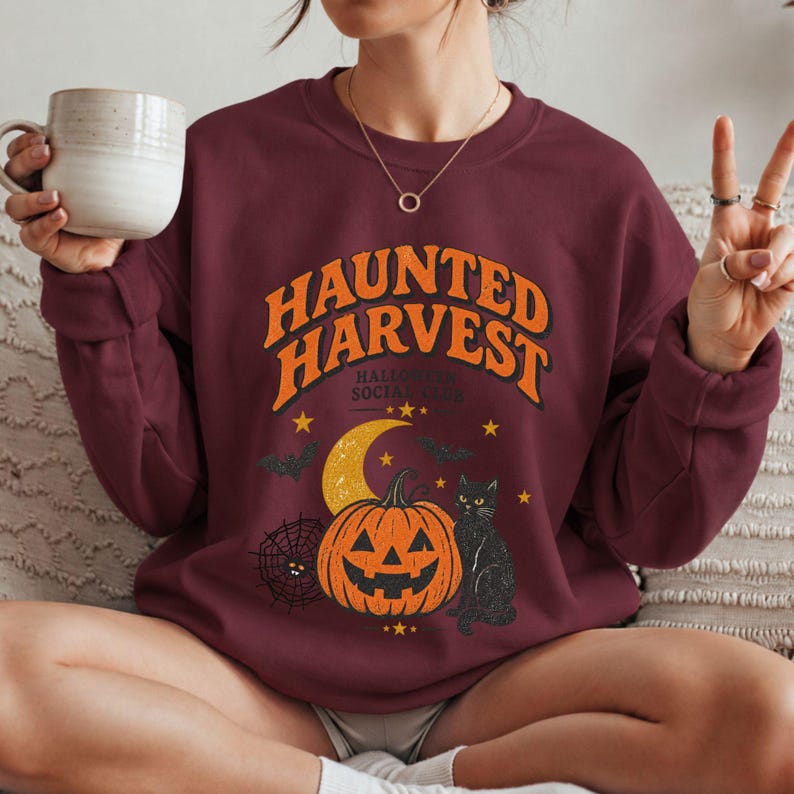 May include: A maroon crewneck sweatshirt featuring the orange text HAUNTED HARVEST and HALLOWEEN SOCIAL CLUB. The graphic includes a yellow crescent moon, a jack-o'-lantern, a black cat, a spider web with a spider, and small bats and stars.