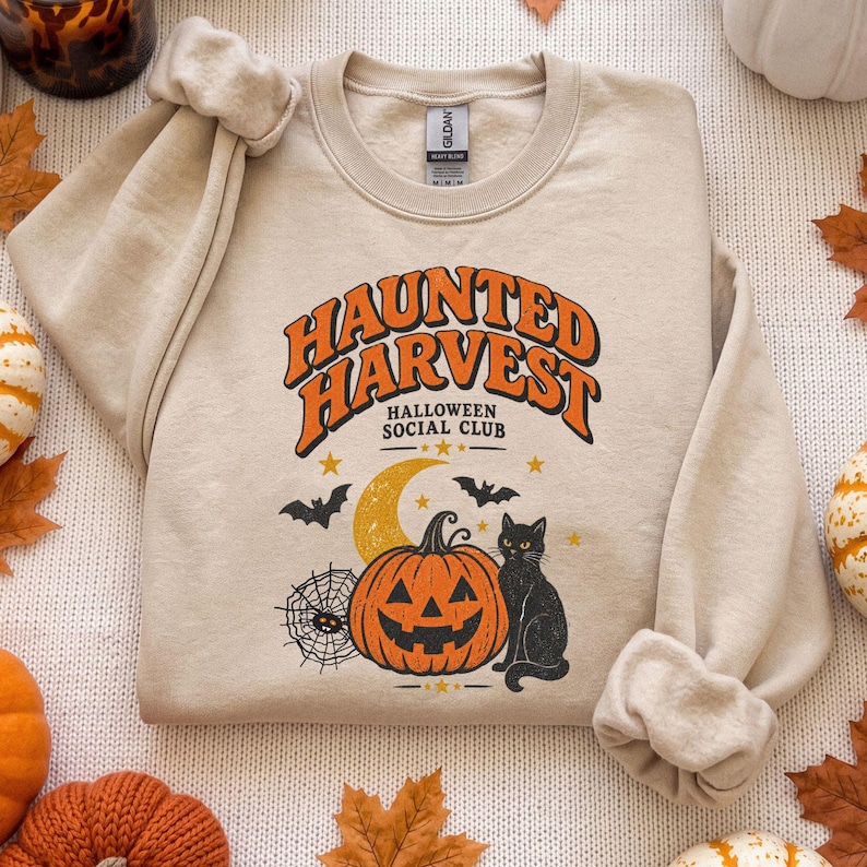May include: A beige crewneck sweatshirt featuring a graphic print with the orange text "HAUNTED HARVEST" and "HALLOWEEN SOCIAL CLUB" above a yellow crescent moon, a jack-o'-lantern, a black cat, a spider web, and small stars.
