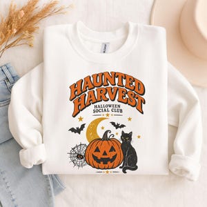 May include: A white crewneck sweatshirt featuring a graphic with the text "HAUNTED HARVEST HALLOWEEN SOCIAL CLUB" in orange and black. The design includes a jack-o'-lantern, a black cat, a crescent moon, a spider web, and flying bats.