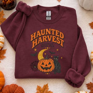 May include: A maroon crewneck sweatshirt featuring a graphic print with the text "HAUNTED HARVEST HALLOWEEN SOCIAL CLUB" in orange. The design includes a yellow crescent moon, an orange jack-o'-lantern, a black cat, and a spider web.