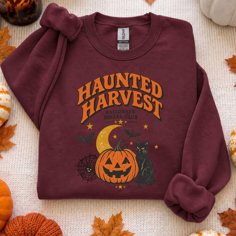 May include: A maroon crewneck sweatshirt featuring a graphic print with the text "HAUNTED HARVEST HALLOWEEN SOCIAL CLUB" in orange. The design includes a yellow crescent moon, an orange jack-o'-lantern, a black cat, and a spider web.