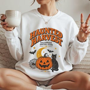 May include: A white crewneck sweatshirt featuring a graphic print with the orange text "HAUNTED HARVEST" and "HALLOWEEN SOCIAL CLUB" above a yellow crescent moon, a black spiderweb, a jack-o'-lantern, a black cat, and small black bats.