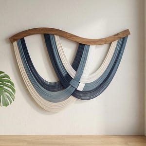 Peut inclure: Une décoration murale composée d'une barre en bois incurvée aux tons naturels, à laquelle sont suspendus plusieurs cordons dans des tons bleu marine, bleu ardoise et crème. Les cordons forment des boucles en U superposées et entrelacées.