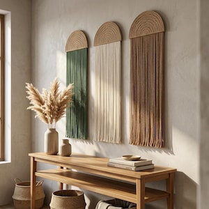 May include: Three semi-circular wall hangings featuring woven tops and long, straight hanging cords in dark green, cream, and brown. Each piece is mounted vertically, showcasing a textured fiber construction with a consistent arched design.