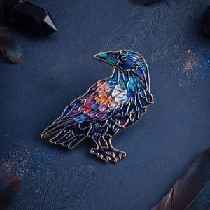 May include: A metal enamel pin shaped like a raven, featuring a stained-glass style design with vibrant segments of blue, purple, orange, and yellow, outlined in gold metal, depicting the bird in profile facing towards the left side.