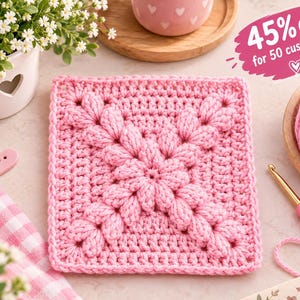 May include: A square pink crochet motif featuring a central puff stitch design. The piece is surrounded by a pink yarn ball, a pink crochet hook, a white flower pot with small white flowers, and a pink heart-shaped button.